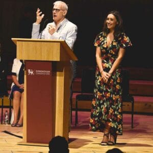 An older man speaks animatedly at a podium while a young woman in a floral dress stands beside him, both on stage in a well-lit auditorium setting.