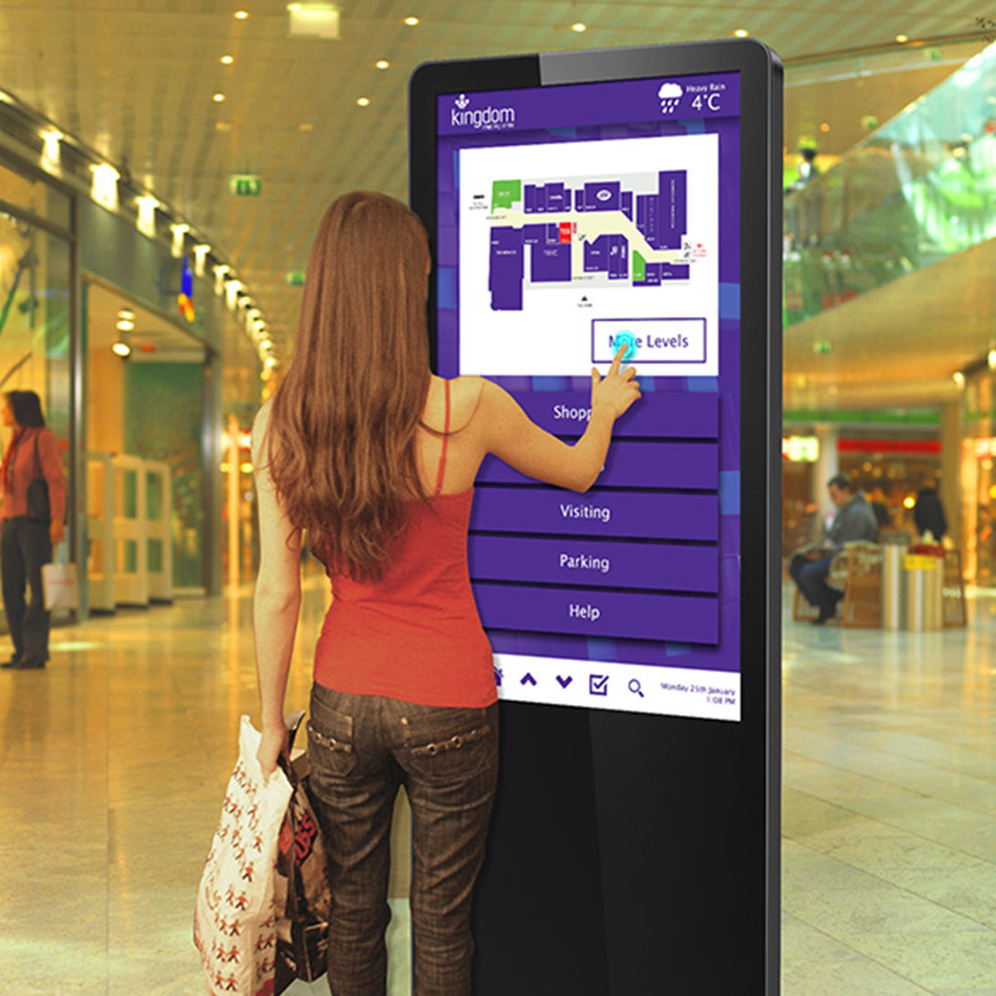 A woman using a black, freestanding interactive digital touch screen totem with wayfinding information on display.