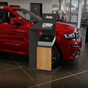 A custom "Jeep" meet and greet station with a digital screen placed in front of a red car in a show room.