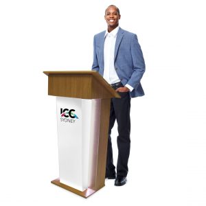 A man in a light blue blazer stands beside a wooden podium with the "ICC Sydney" logo, smiling confidently with one hand in his pocket against a plain white background.