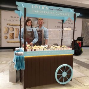 A mobile food cart with a woman and a man stood behind, with some hand crafted cupcakes behind a bespoke food screen.