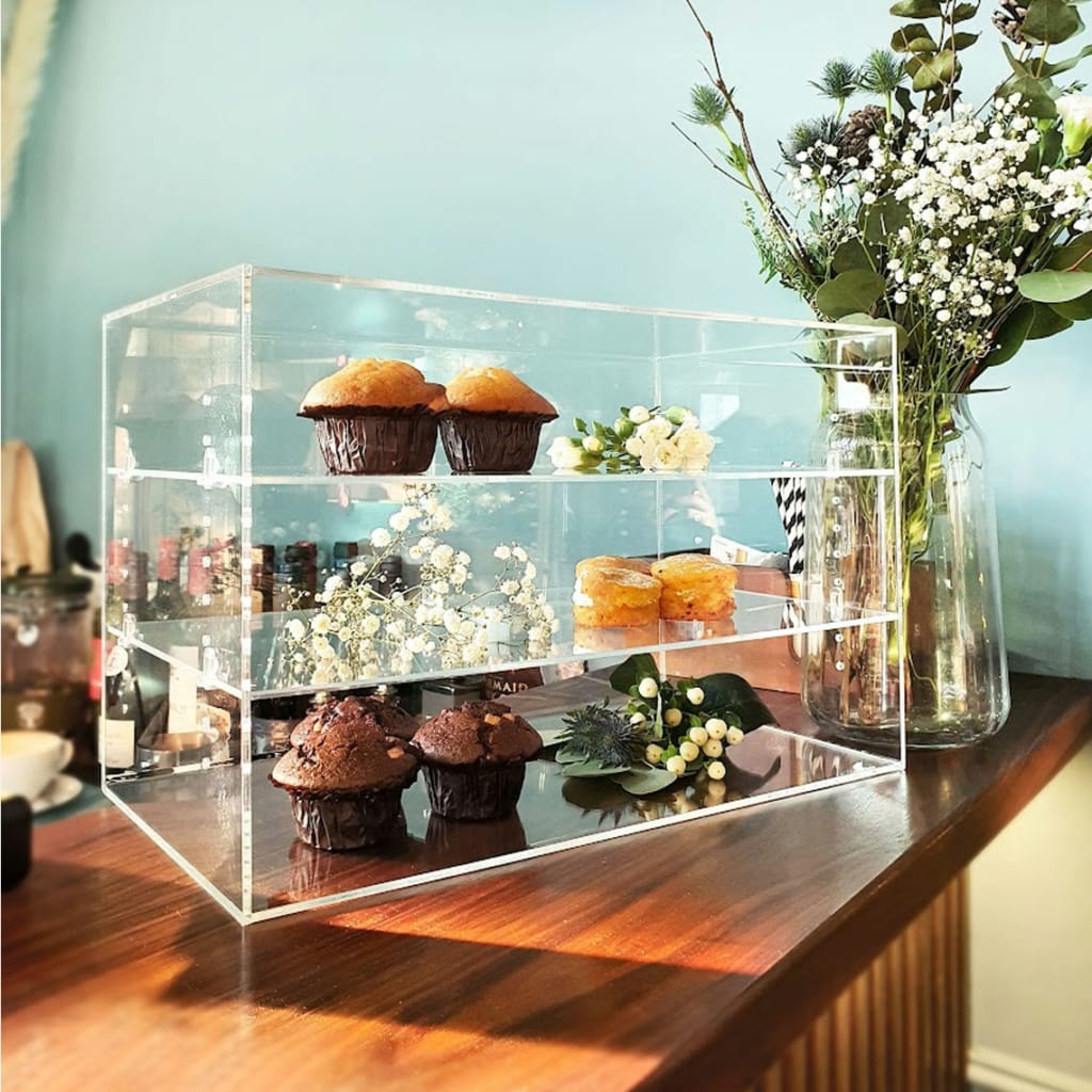 A clear Acrylic Display Unit with three shelves on a front shop counter, holding a selection of cakes and decorative flowers.